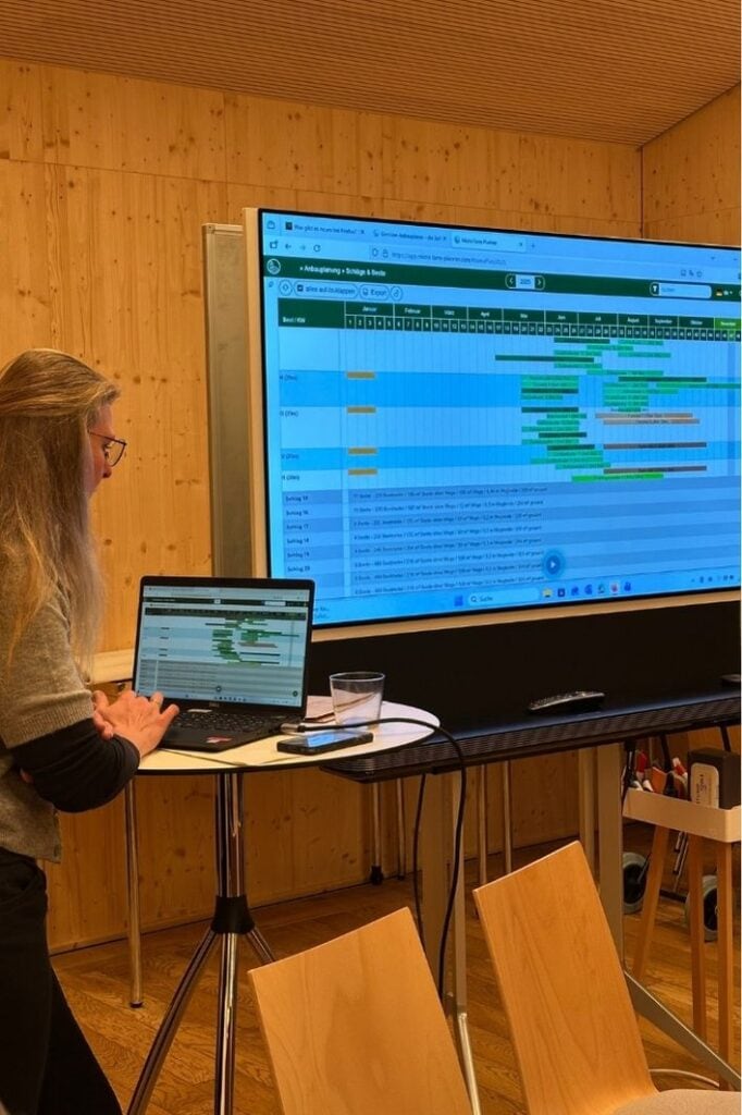 Mitgliederversammlung Selbsterntegarten. Man sieht eine Frau an einem Laptop und einen großen Bildschrim mit einem Diagramm in einem Raum mit viel Holz